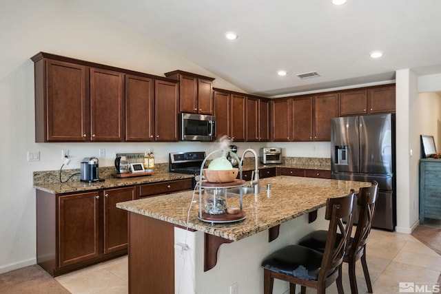 kitchen with a kitchen island with sink, vaulted ceiling, light tile patterned floors, light stone countertops, and appliances with stainless steel finishes