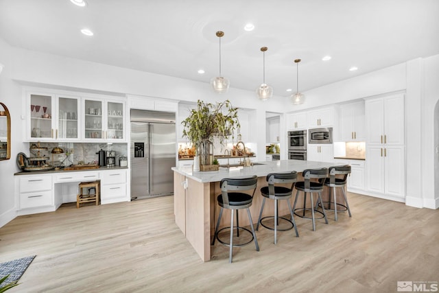 kitchen featuring pendant lighting, a kitchen island with sink, built in appliances, light hardwood / wood-style floors, and white cabinetry