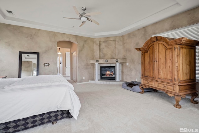 bedroom featuring ceiling fan, a tile fireplace, light carpet, and a tray ceiling