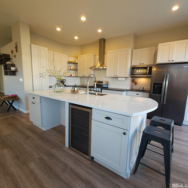 kitchen with an island with sink, wall chimney range hood, beverage cooler, white cabinetry, and appliances with stainless steel finishes