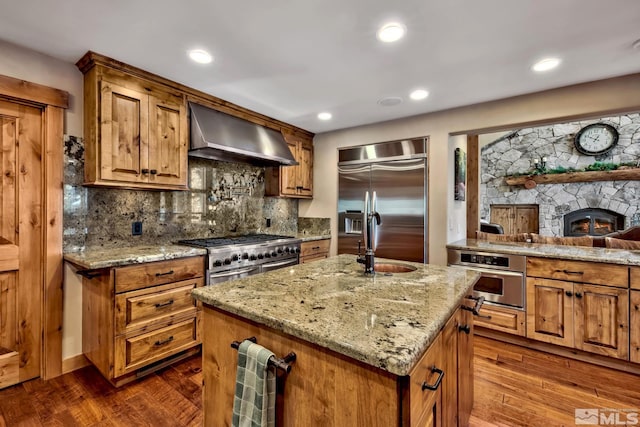 kitchen with light stone counters, wall chimney exhaust hood, hardwood / wood-style flooring, a center island with sink, and high quality appliances