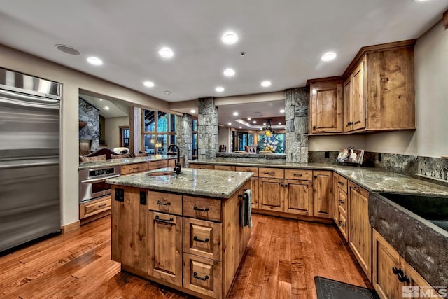 kitchen with stainless steel appliances, an island with sink, light wood-type flooring, stone counters, and sink