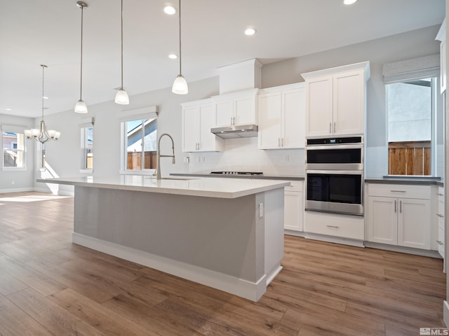 kitchen featuring white cabinets, decorative light fixtures, stainless steel double oven, and a center island with sink