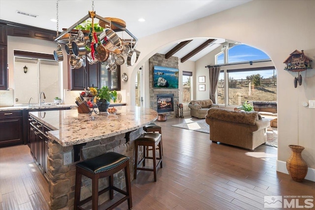 kitchen with a stone fireplace, decorative backsplash, a kitchen island, vaulted ceiling with beams, and dark brown cabinetry