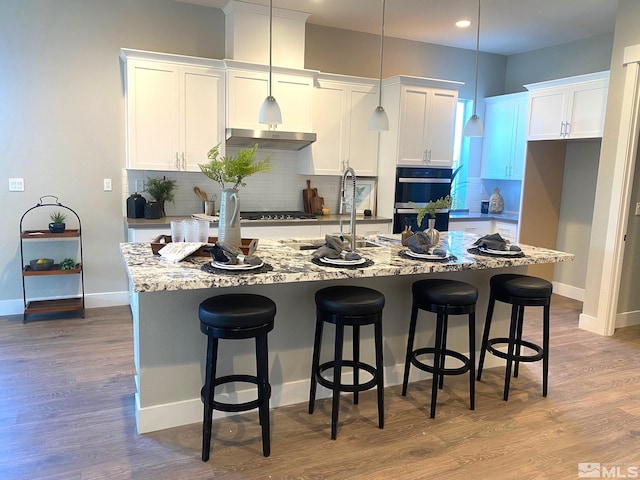 kitchen with light stone countertops, decorative light fixtures, decorative backsplash, a kitchen island with sink, and white cabinetry