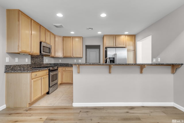 kitchen featuring stainless steel appliances, light brown cabinetry, backsplash, dark stone counters, and a kitchen breakfast bar