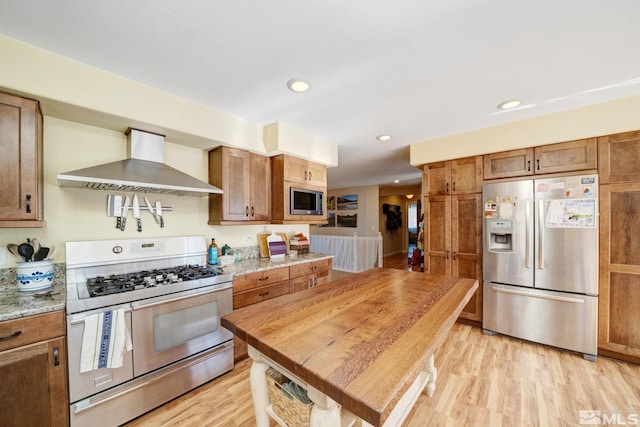 kitchen with light stone counters, appliances with stainless steel finishes, wall chimney range hood, and light hardwood / wood-style floors