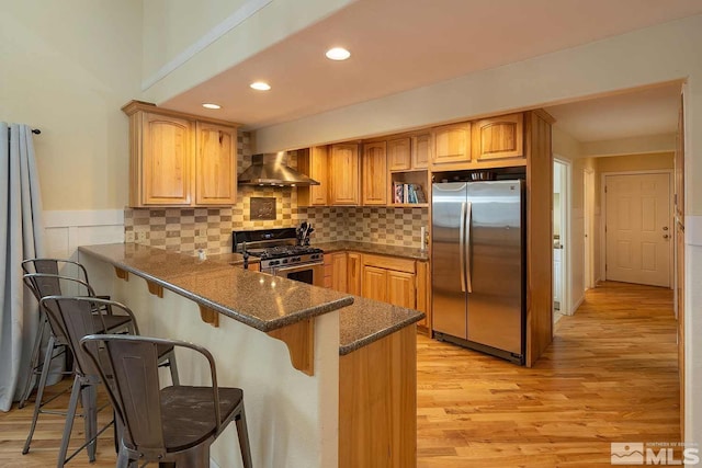 kitchen with kitchen peninsula, decorative backsplash, dark stone counters, wall chimney range hood, and appliances with stainless steel finishes