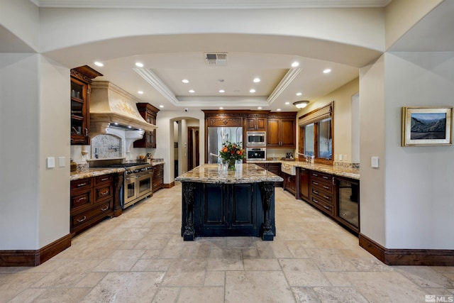 kitchen featuring light stone countertops, premium range hood, a raised ceiling, a kitchen island with sink, and appliances with stainless steel finishes