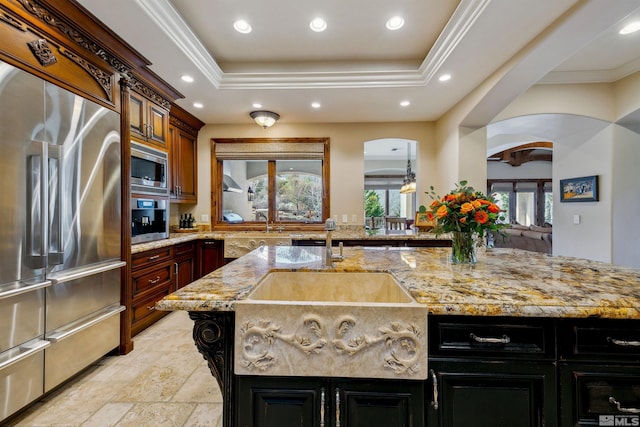 kitchen with light stone countertops, a tray ceiling, an island with sink, and appliances with stainless steel finishes