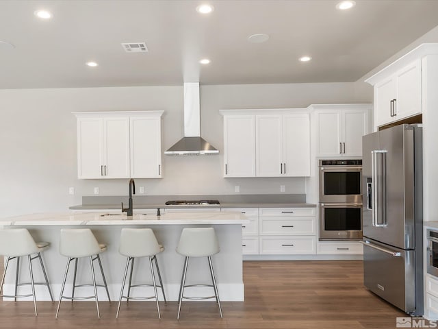 kitchen featuring stainless steel appliances, a kitchen bar, sink, white cabinetry, and wall chimney range hood