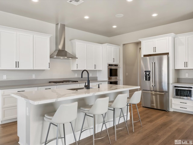 kitchen featuring wall chimney exhaust hood, white cabinets, a kitchen island with sink, and appliances with stainless steel finishes