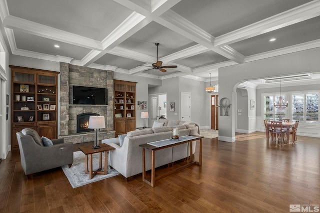 living room with beam ceiling, crown molding, a stone fireplace, built in shelves, and ceiling fan with notable chandelier