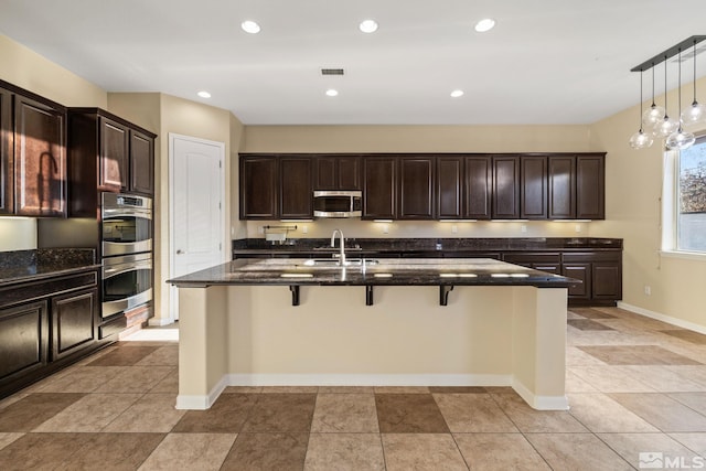 kitchen with sink, hanging light fixtures, a kitchen island with sink, stainless steel appliances, and dark stone counters