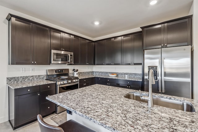 kitchen featuring stainless steel appliances, recessed lighting, dark brown cabinetry, a sink, and light stone countertops