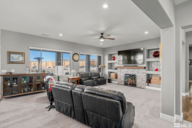 living room with ceiling fan, light colored carpet, a stone fireplace, and built in shelves