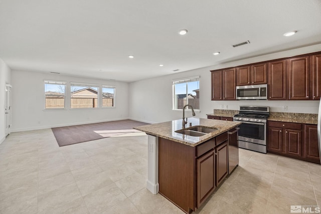 kitchen with sink, dark brown cabinets, stainless steel appliances, light stone counters, and a center island with sink