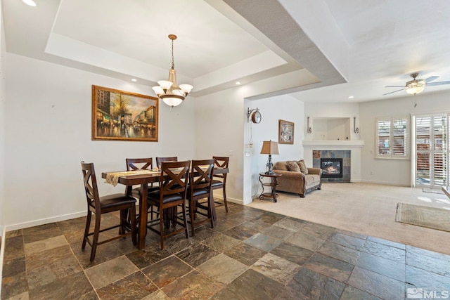 dining room with a tiled fireplace, ceiling fan with notable chandelier, dark carpet, and a tray ceiling