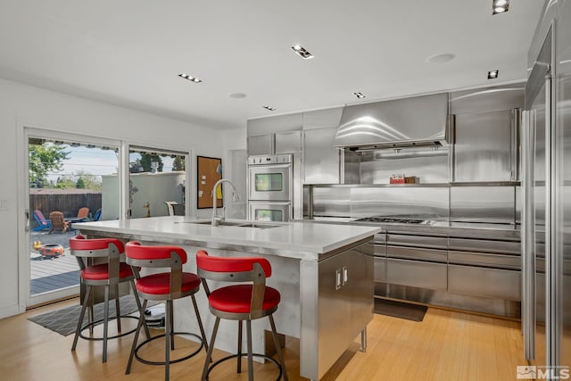 kitchen featuring light hardwood / wood-style flooring, sink, exhaust hood, a kitchen island with sink, and a breakfast bar