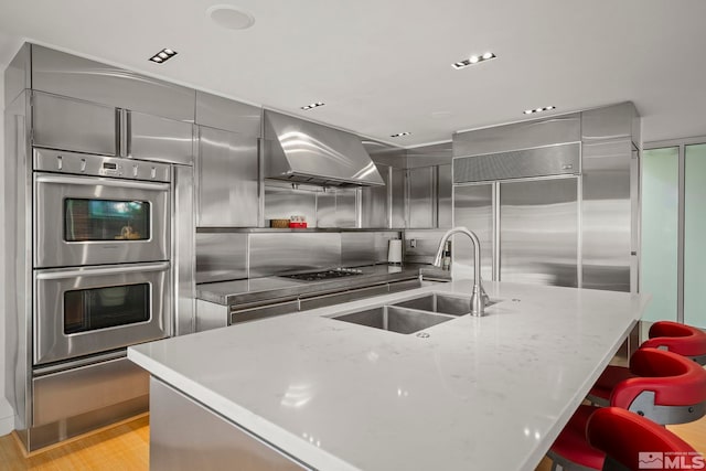 kitchen with light stone countertops, stainless steel appliances, an island with sink, sink, and ventilation hood