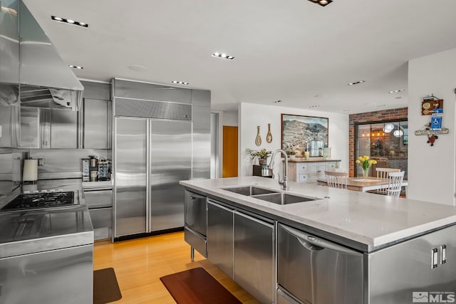 kitchen with appliances with stainless steel finishes, island exhaust hood, sink, light wood-type flooring, and a center island with sink