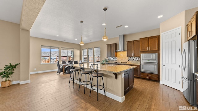 kitchen featuring hanging light fixtures, appliances with stainless steel finishes, a kitchen breakfast bar, a kitchen island with sink, and wall chimney range hood