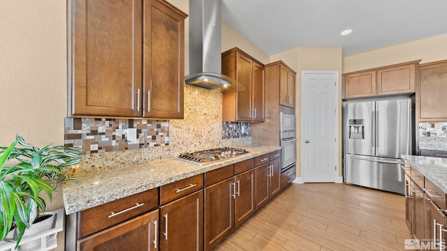 kitchen with backsplash, light stone counters, stainless steel appliances, wall chimney range hood, and light wood-type flooring