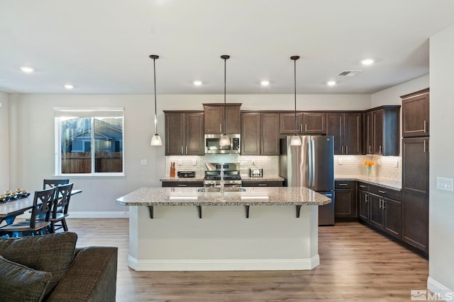 kitchen with pendant lighting, a breakfast bar area, appliances with stainless steel finishes, a kitchen island with sink, and light stone countertops