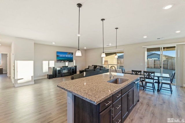 kitchen with an island with sink, sink, dark brown cabinetry, light stone countertops, and light wood-type flooring