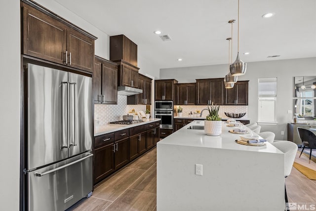 kitchen with dark brown cabinetry, hanging light fixtures, an island with sink, stainless steel appliances, and backsplash