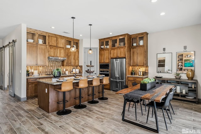 kitchen with a kitchen island with sink, hanging light fixtures, stainless steel appliances, light stone counters, and a barn door