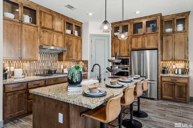 kitchen featuring light stone counters, tasteful backsplash, decorative light fixtures, a center island with sink, and appliances with stainless steel finishes