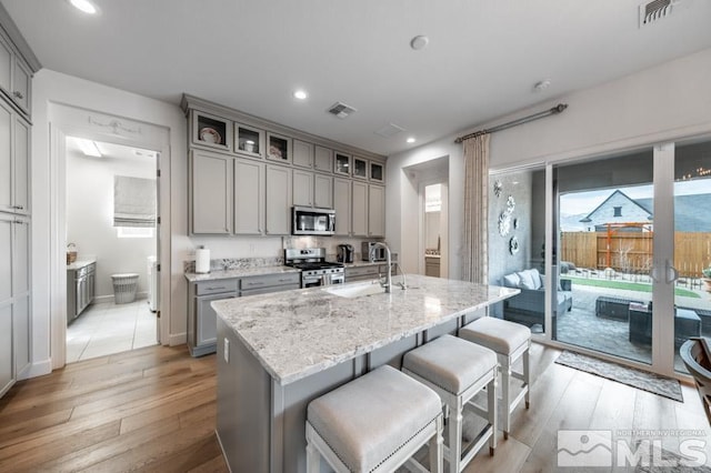 kitchen with gray cabinetry, light stone counters, light hardwood / wood-style flooring, an island with sink, and stainless steel appliances