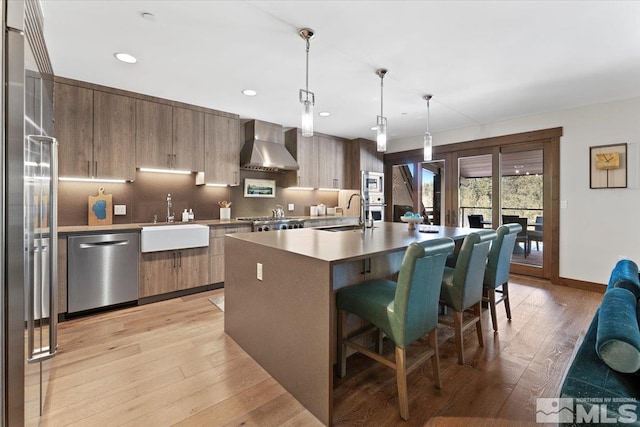 kitchen with wall chimney exhaust hood, sink, decorative light fixtures, appliances with stainless steel finishes, and an island with sink