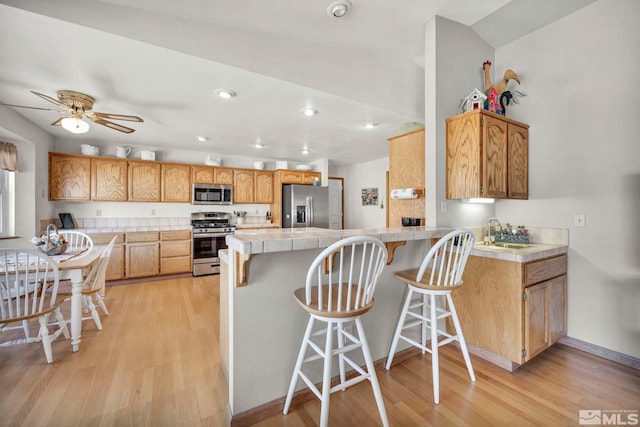 kitchen with tile countertops, light wood-type flooring, appliances with stainless steel finishes, kitchen peninsula, and ceiling fan