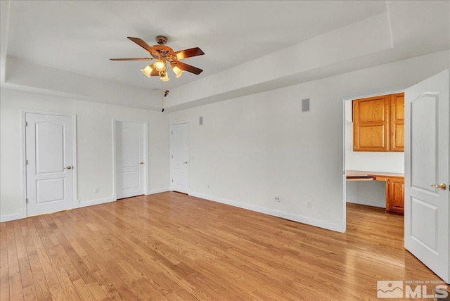 unfurnished room with a raised ceiling, built in desk, ceiling fan, and light hardwood / wood-style flooring