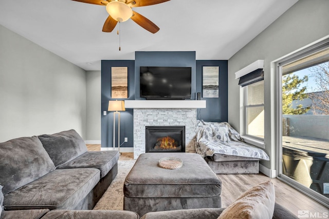 living area with baseboards, a stone fireplace, wood finished floors, and a ceiling fan