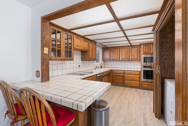 kitchen with tile counters, kitchen peninsula, stainless steel appliances, light hardwood / wood-style floors, and decorative backsplash