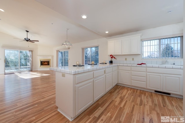 kitchen with tile countertops, light wood-type flooring, kitchen peninsula, pendant lighting, and white cabinets