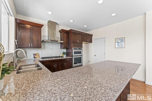 kitchen featuring sink, wall chimney range hood, stainless steel appliances, light stone countertops, and backsplash