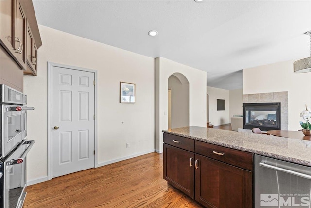 kitchen with dark brown cabinetry, light stone counters, light wood-type flooring, stainless steel appliances, and a tiled fireplace