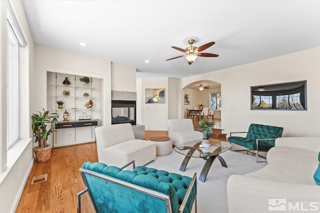 living room featuring a multi sided fireplace, built in shelves, ceiling fan, and light hardwood / wood-style flooring