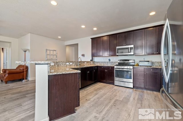 kitchen featuring dark brown cabinets, light wood-type flooring, kitchen peninsula, stainless steel appliances, and light stone countertops