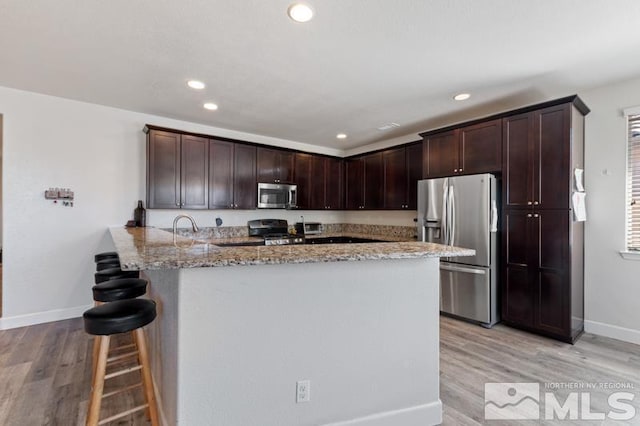 kitchen featuring appliances with stainless steel finishes, kitchen peninsula, light stone countertops, dark brown cabinets, and light hardwood / wood-style flooring