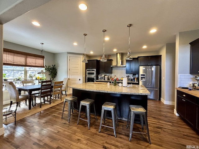 kitchen with appliances with stainless steel finishes, decorative light fixtures, dark hardwood / wood-style flooring, a kitchen island with sink, and wall chimney exhaust hood