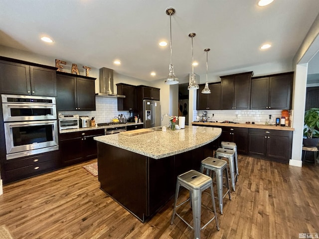 kitchen with dark brown cabinetry, wall chimney exhaust hood, appliances with stainless steel finishes, pendant lighting, and a kitchen island with sink