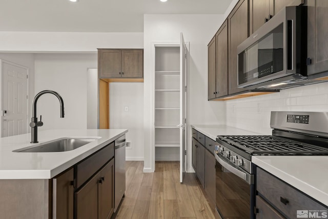 kitchen featuring an island with sink, sink, decorative backsplash, stainless steel appliances, and light wood-type flooring