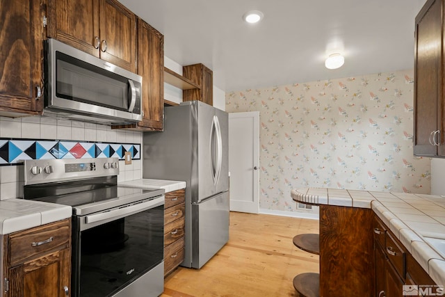 kitchen with tile countertops, backsplash, light hardwood / wood-style floors, stainless steel appliances, and dark brown cabinets