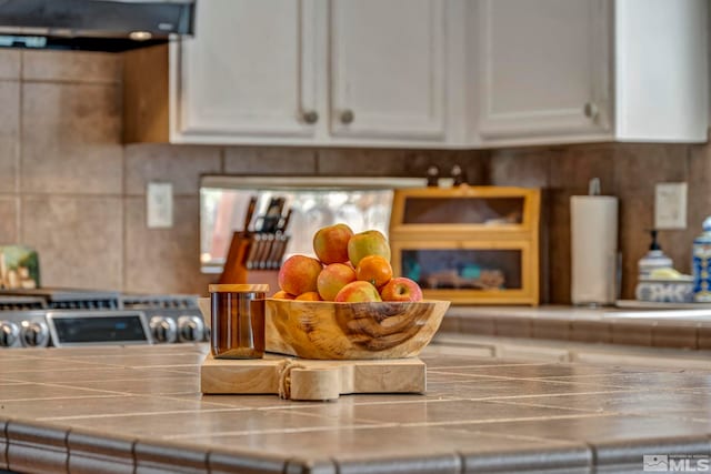 interior details with tasteful backsplash, tile countertops, exhaust hood, and white cabinets