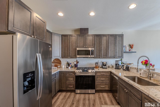 kitchen featuring sink, appliances with stainless steel finishes, dark brown cabinets, light stone countertops, and light wood-type flooring